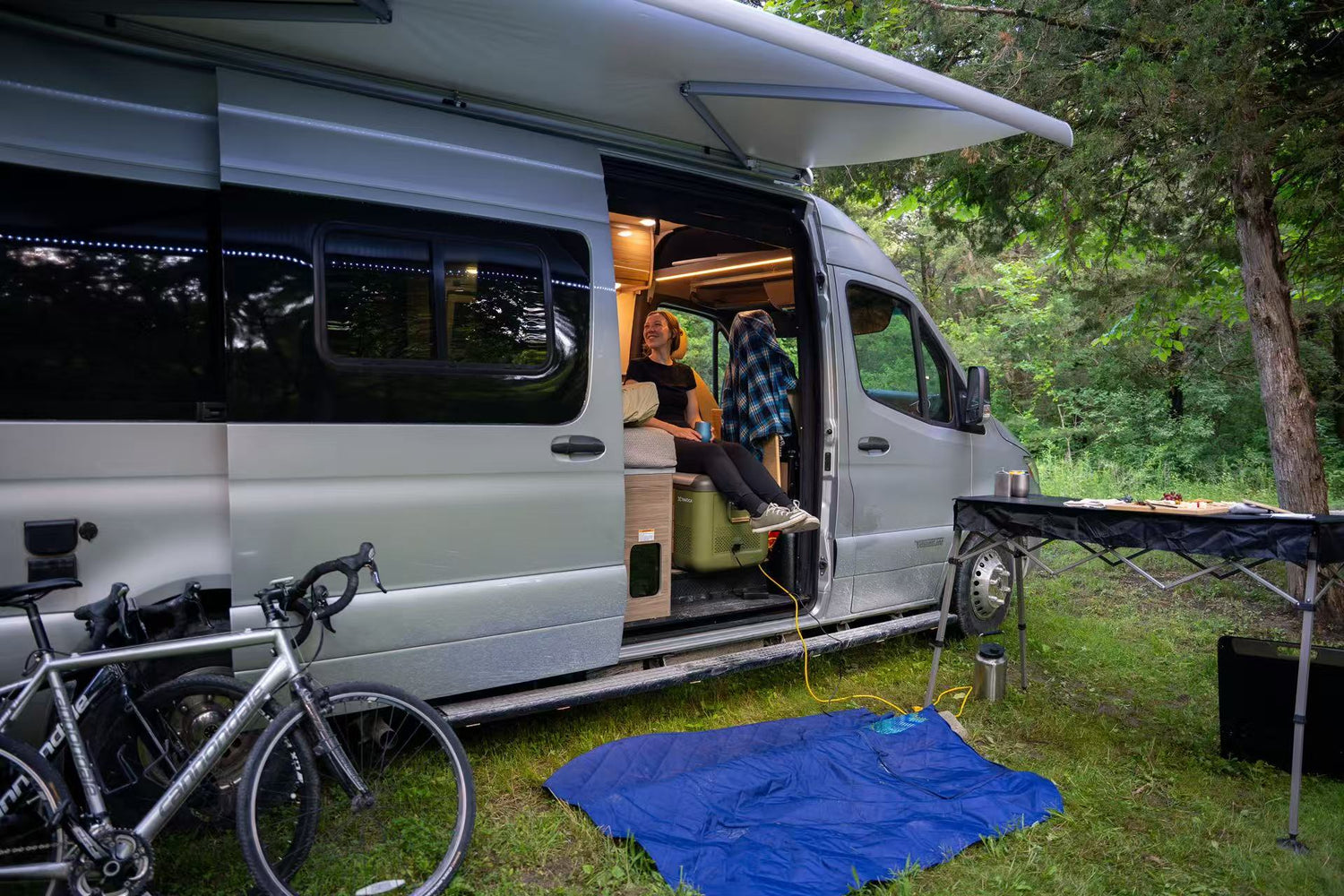 TRAVOCA portable car fridge placed inside an RV during outdoor camping, with a woman sitting on it while enjoying the van life atmosphere.
